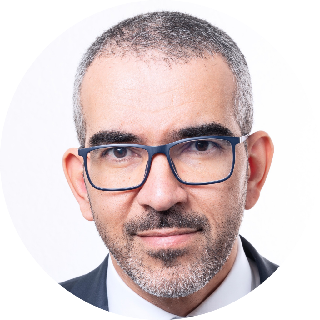 Close-up portrait of a man with salt-and-pepper hair and blue-framed glasses, wearing a suit, against a white background in a circular crop.